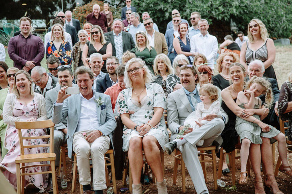 Wedding guests laugh together, with phones raised to photograph the ceremony.