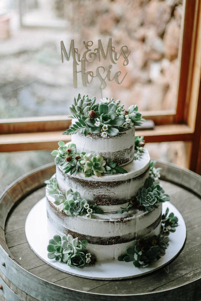 A three-tier semi-naked cake decorated with succulents stands on a barrel at Camp Sunnystones.
