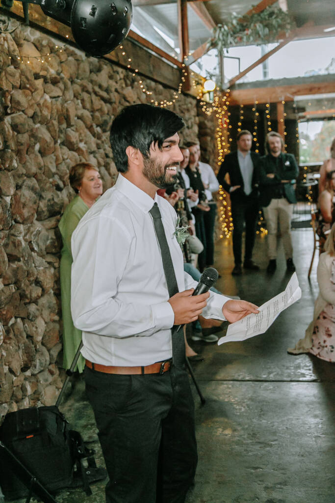A wedding attendant reads a speech beside the stone wall and fairy lights at Camp Sunnystones.