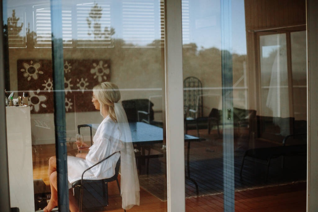 Through glass doors, the bride sits in a satin robe with her veil pinned, flute in hand.