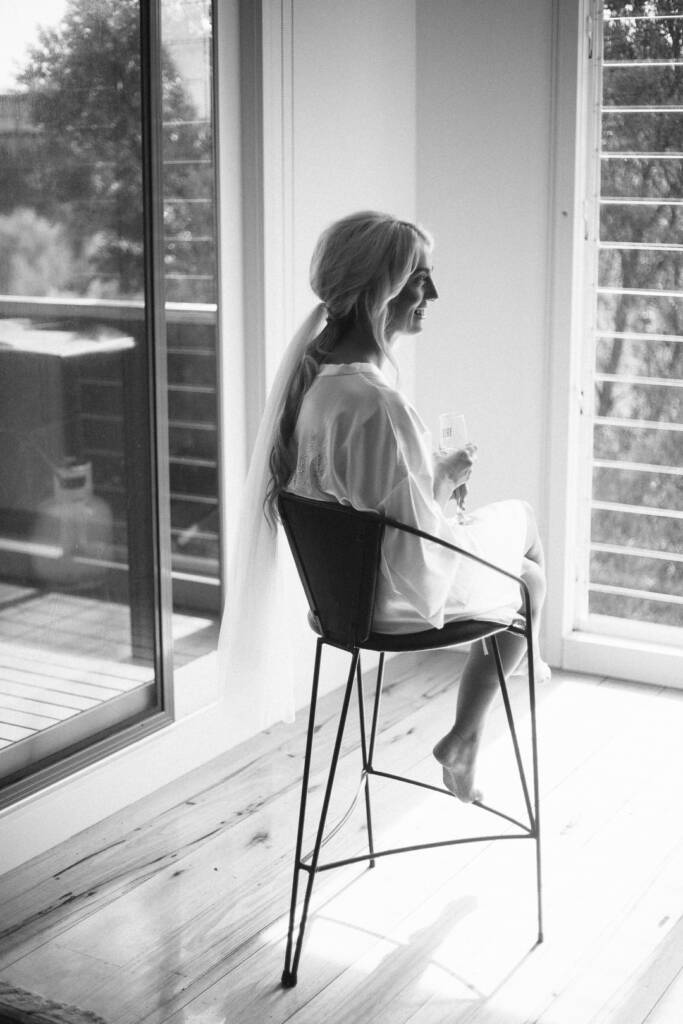 Black and white of the bride laughing on a wire stool by louvred windows, robe and veil on.