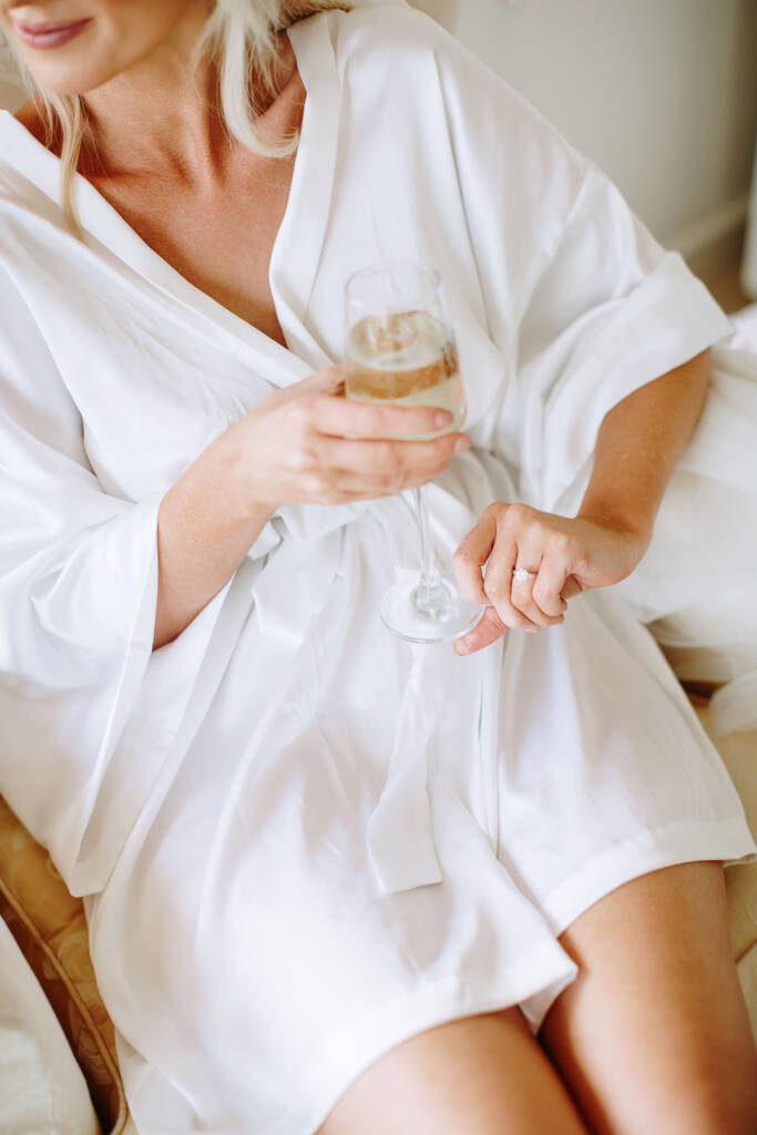 Close on the bride in her robe looking down at a champagne flute, ring catching the light.
