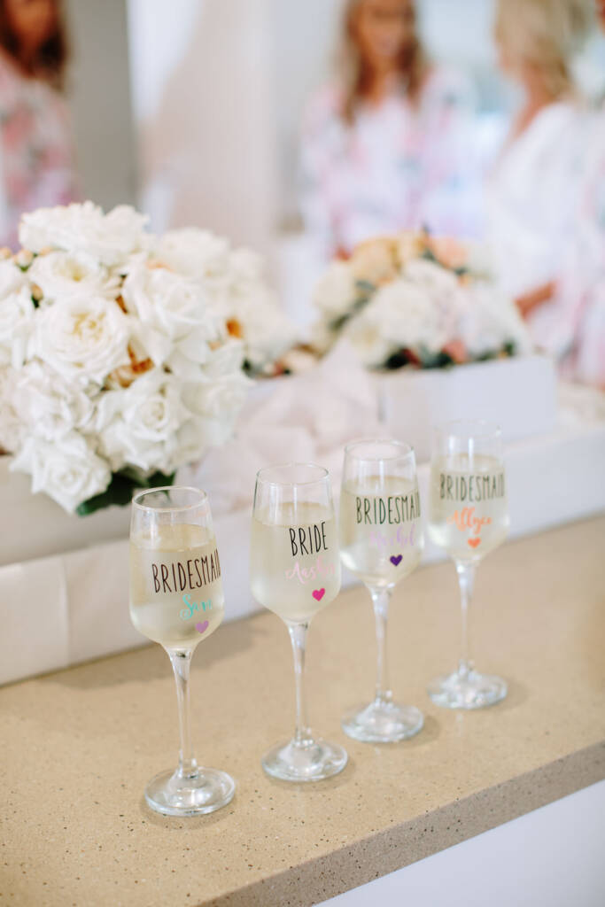 Four lettered champagne flutes in a row on a stone bench, white roses and a peach bouquet behind.