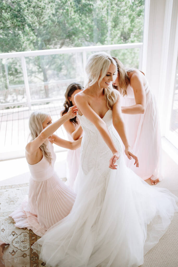 Three bridesmaids in blush pleated gowns fasten Aasher into the lace gown by a bright window.