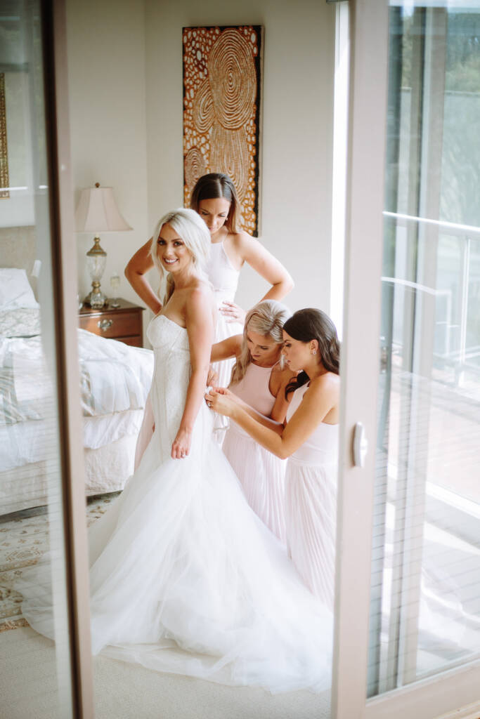 Seen through a doorway, Aasher stands hand on hip in the full gown as bridesmaids fasten her.