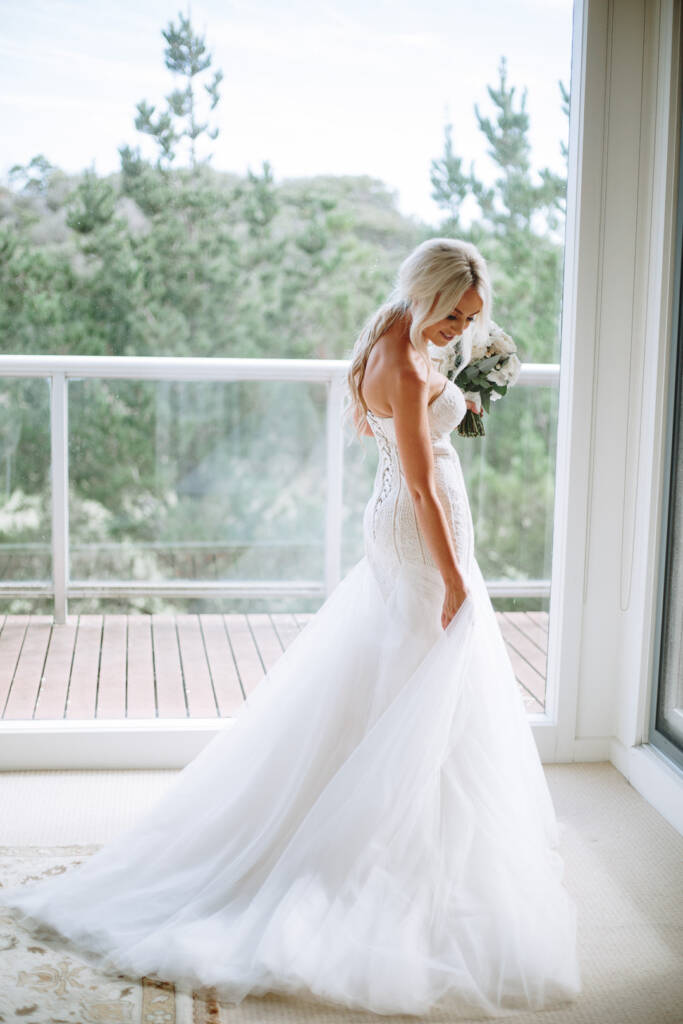 Aasher lifts the tulle skirt with one hand at the balcony doors, bouquet in the other.