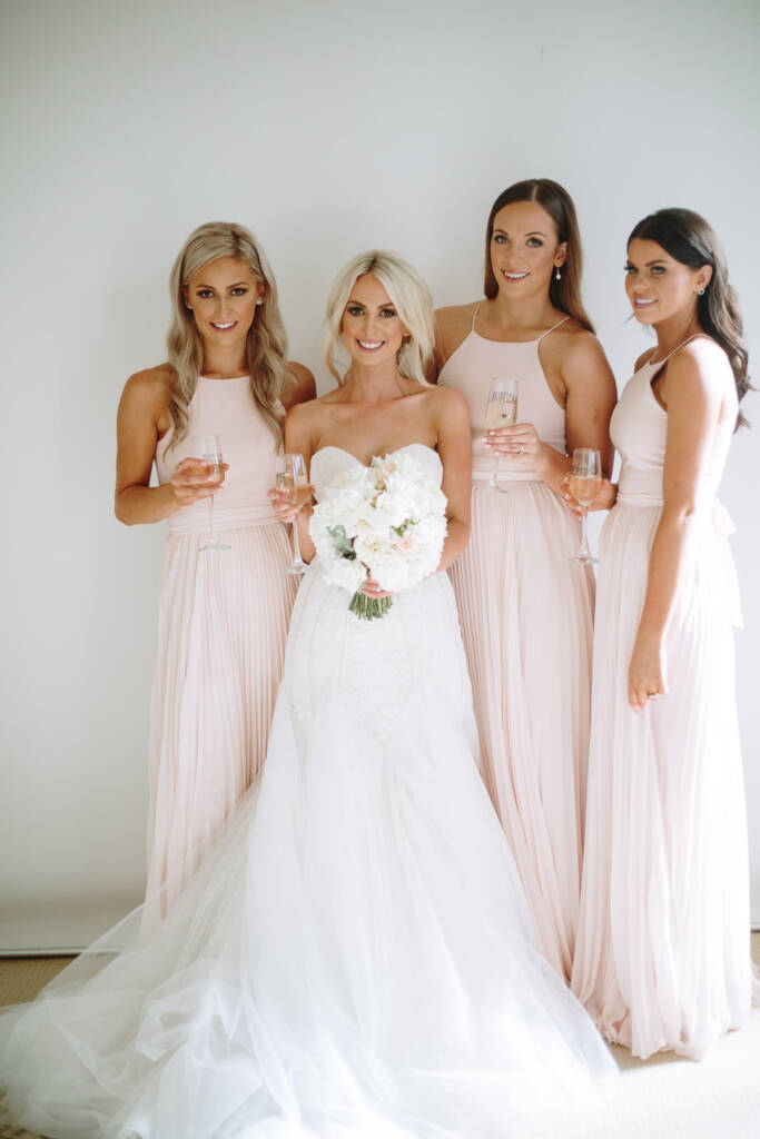 Bridal party portrait of Aasher with three bridesmaids in blush halter gowns, flutes in hand.