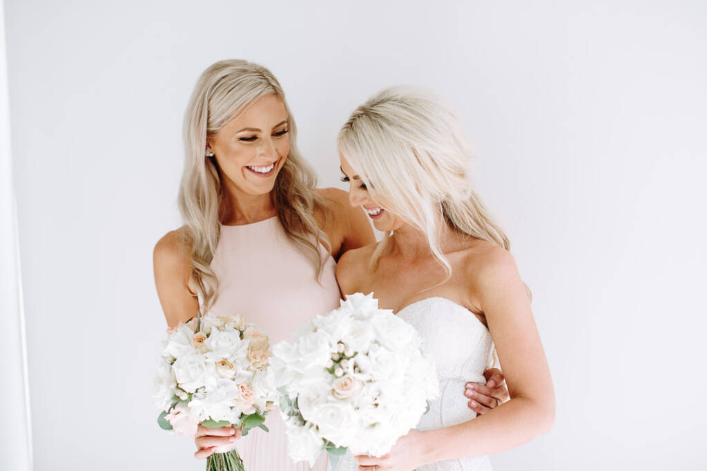 Aasher and a bridesmaid laugh together holding white and peach bouquets against a white wall.
