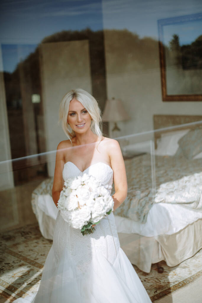 Aasher smiles with her bouquet behind glass at her Cape Schanck Airbnb, trees reflected around her.