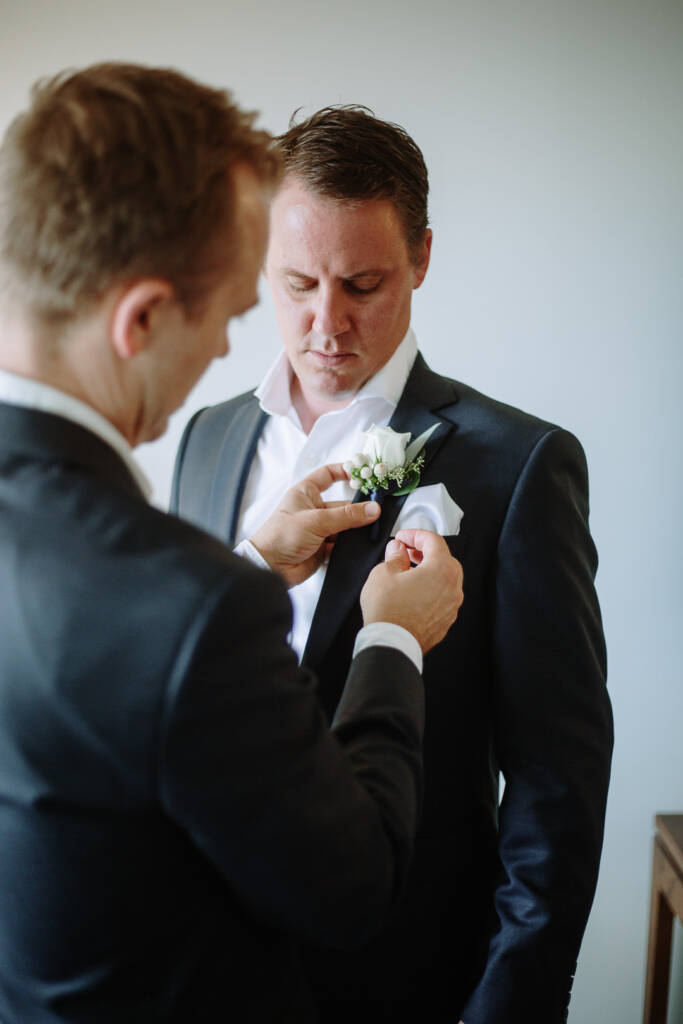 A groomsman pins a white rose and berry boutonniere to Nick's charcoal lapel.