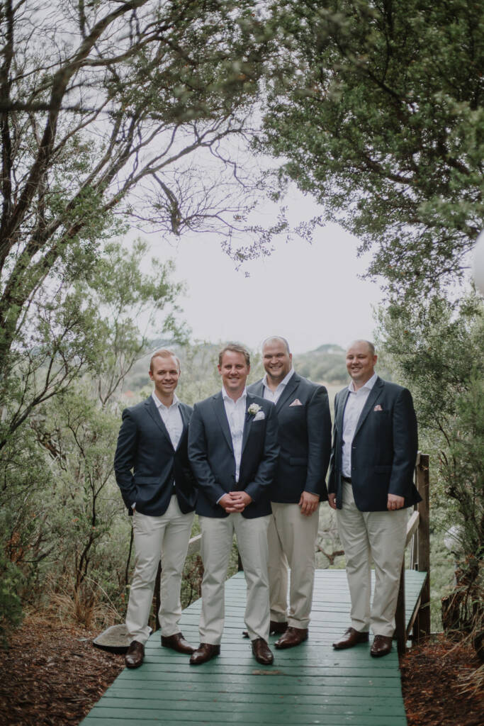 Nick and three groomsmen line a green boardwalk beneath an arch of moonah and tea tree.