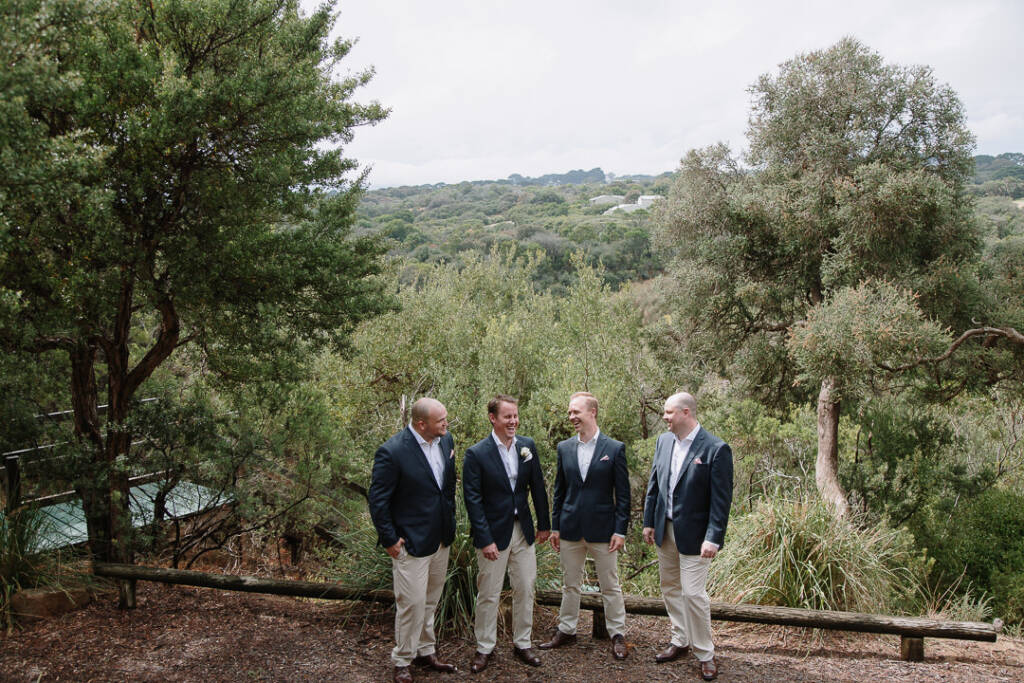 Four men laugh in a row beside a log bench on a rise above dense coastal scrub.