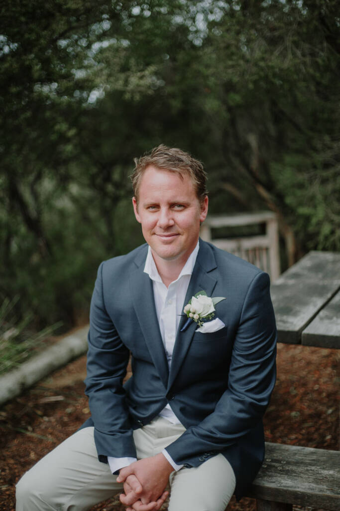 Groom portrait of Nick on a timber picnic bench, white rose boutonniere, moonah trees behind.