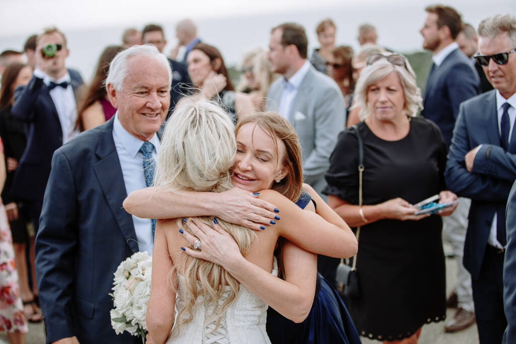 From behind, Aasher's laced bodice as she hugs a woman in navy who smiles with eyes closed.