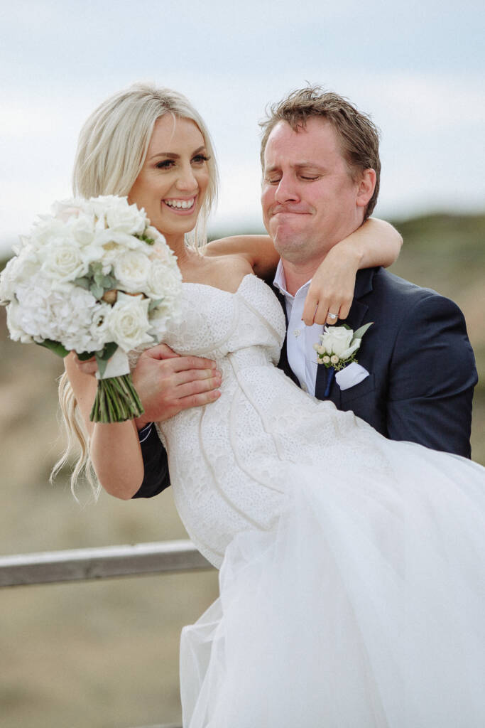 Nick carries Aasher beside the lookout above Fingal Beach, laughing as she holds her bouquet.