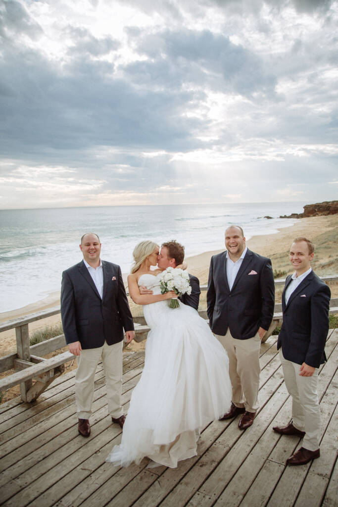 Aasher and Nick kiss on the boardwalk above Fingal Beach, three bridesmaids beside them.