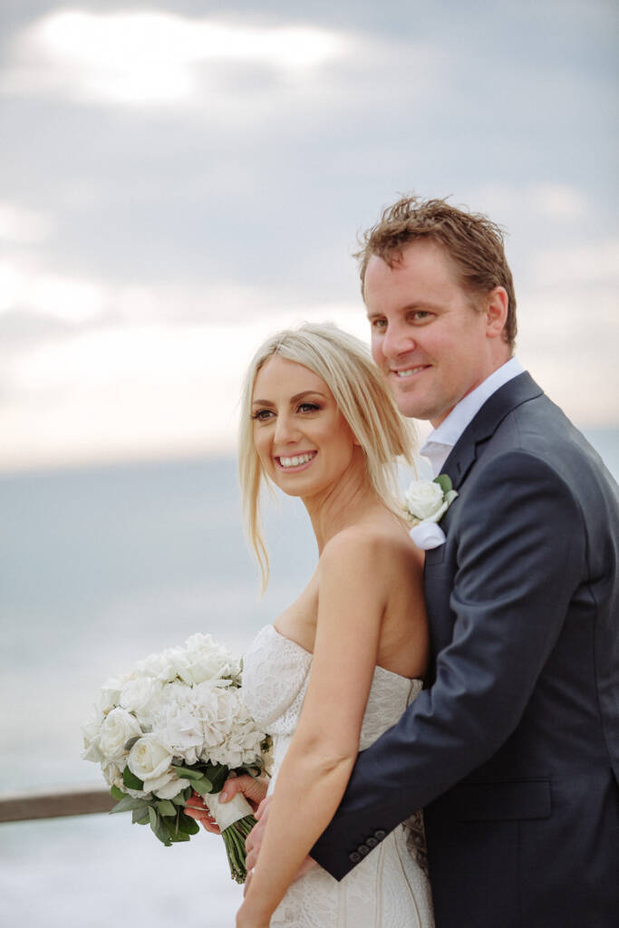 Aasher smiles with her white bouquet as Nick leans close at Fingal Beach lookout.