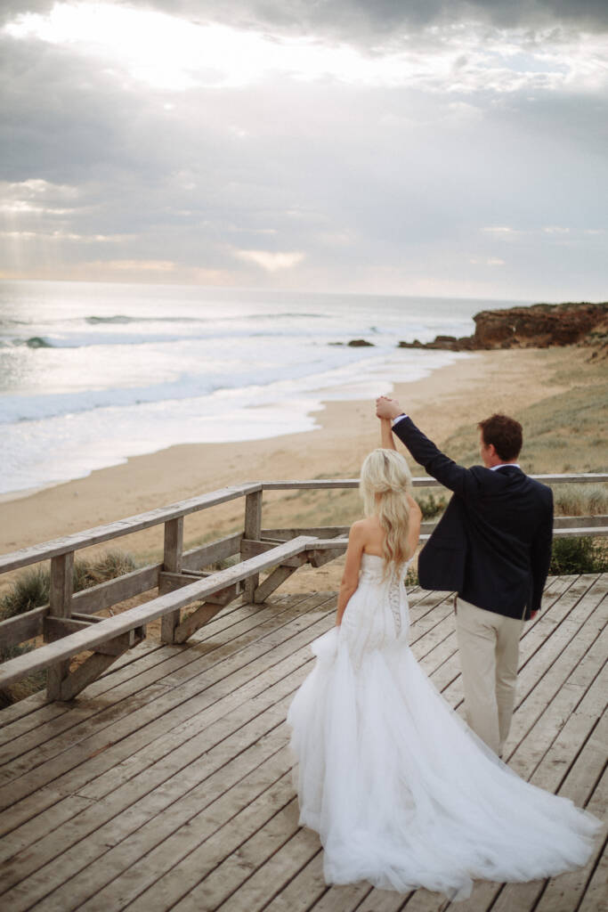 Nick raises their joined hands at the boardwalk rail above Fingal Beach and its headland.