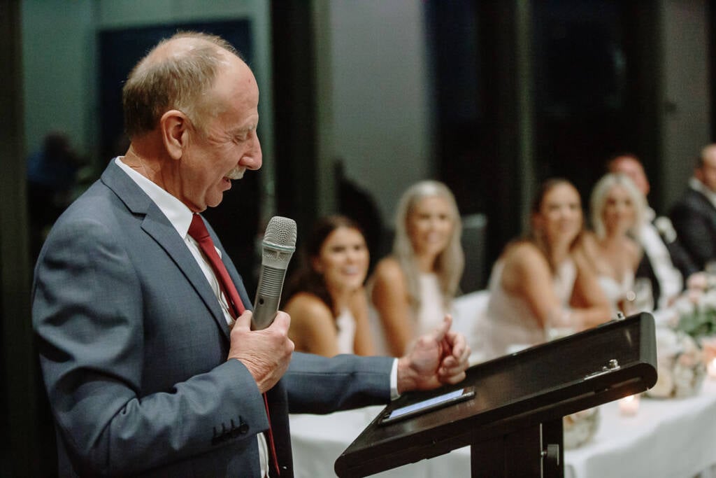 A speaker in a grey suit reads from a tablet at the lectern, the bridal party behind.