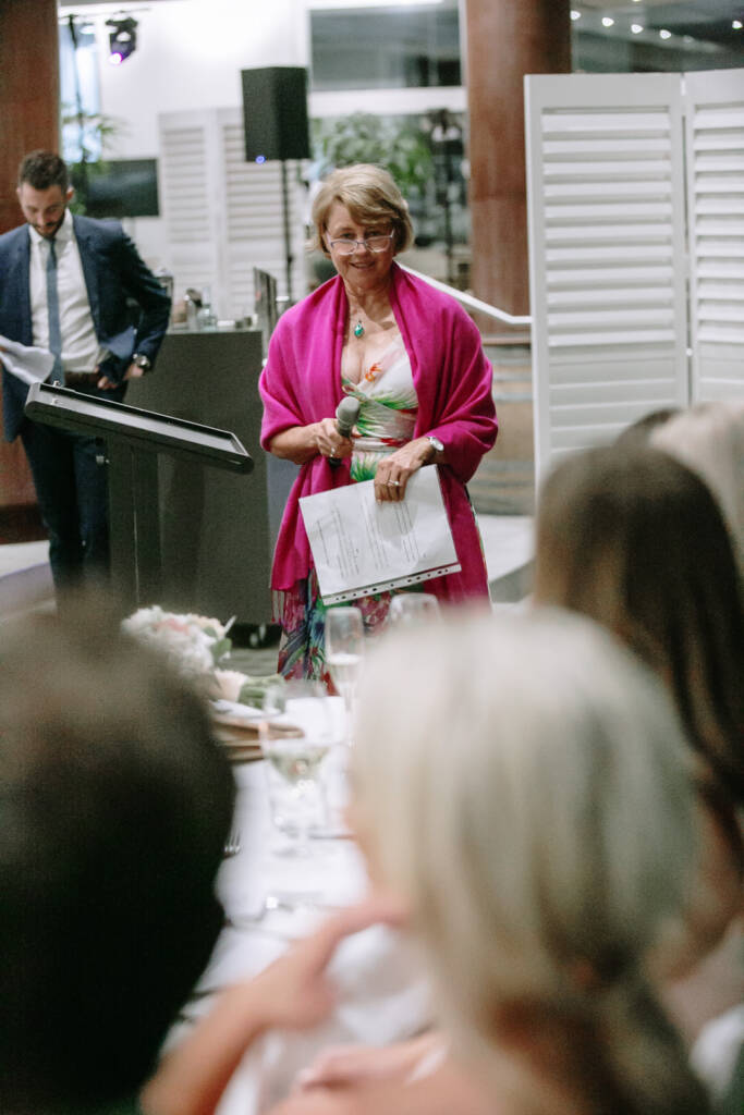 A speaker in a magenta wrap holds the microphone and a printed page before the lectern.