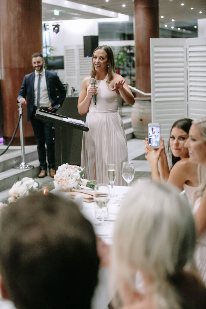 A bridesmaid in a blush pleated gown gives her speech at the lectern, gesturing to the room.