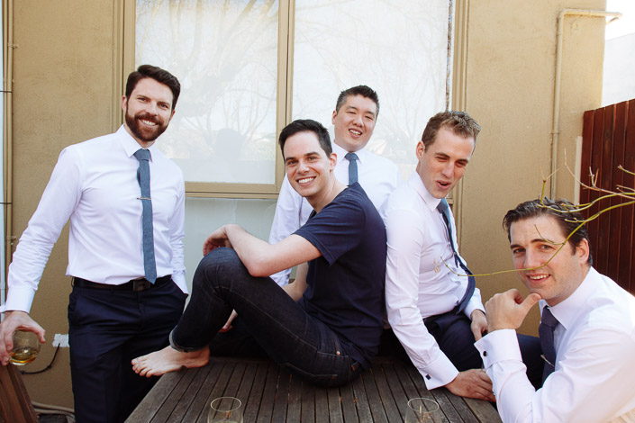 Five men gather beside a wooden table, one seated barefoot.