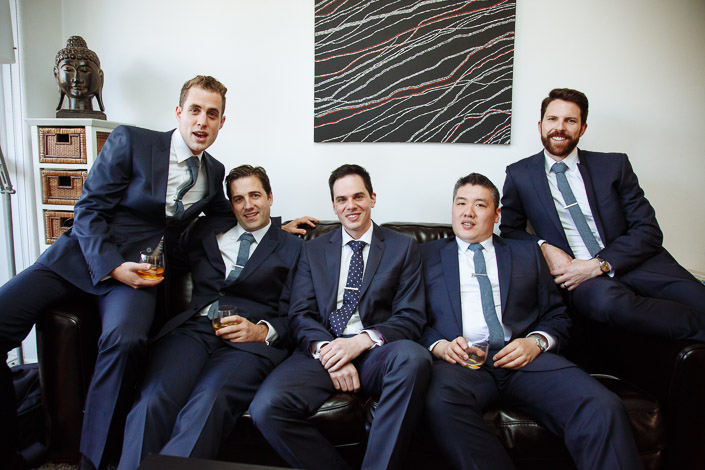Five men in navy suits sit together holding drinks at the groom's home.