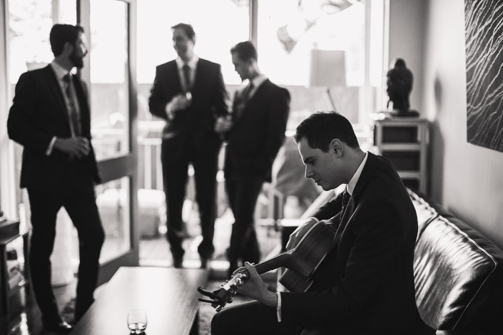 A man plays an acoustic guitar while three groomsmen stand near glass doors.