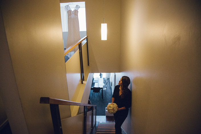 Pascale holds her bouquet on the stairs at Quest on William, looking towards her hanging wedding dress.