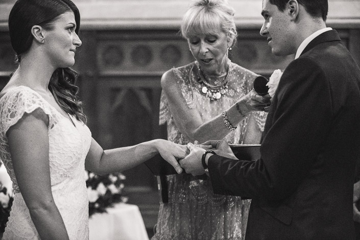 The bride's hand rests in the groom's for the ring, in black and white.