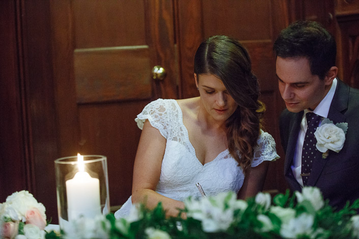 Pascale signs the marriage register beside Pasquale, with flowers and a candle on the table at Chapter House.