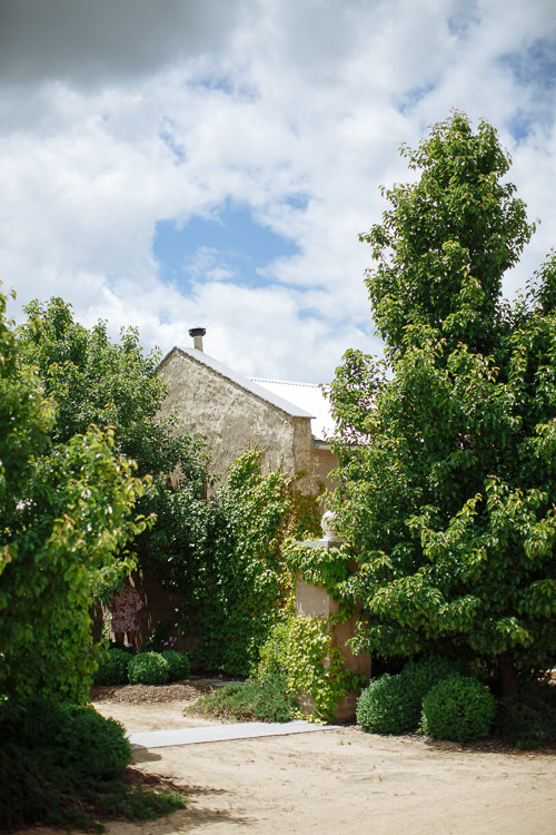 The gable of a rendered stone building rises above pear trees and clipped box balls.