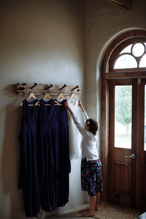 A girl with a flower in her hair reaches up to a peg rail of navy bridesmaid dresses.