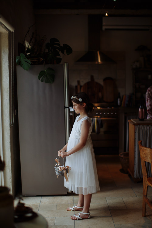 A flower girl in white tulle and a flower crown holds her petal basket in the kitchen.