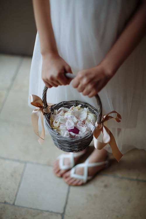 The flower girl's hands hold a grey wicker basket of pink petals tied with peach ribbon.