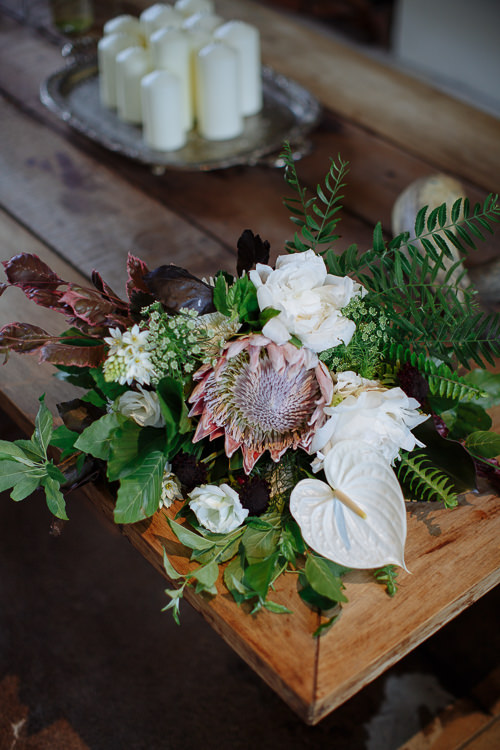 A bouquet of king protea, white roses, anthurium and ferns lies on a timber table beside pillar candles.