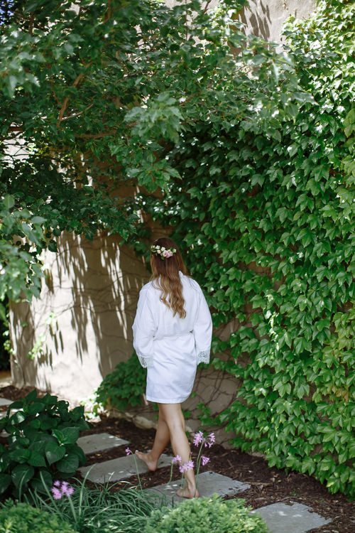 From behind, Elysha in a white robe and flower crown walks barefoot along stepping stones beneath ivy.