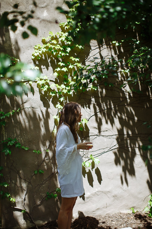 Elysha in her white robe holds a glass of champagne against an ivy shadowed rendered wall.