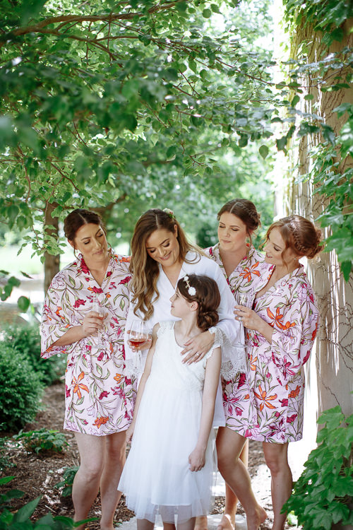 Three bridesmaids in pink floral robes and Elysha gather around the flower girl beneath a tree.
