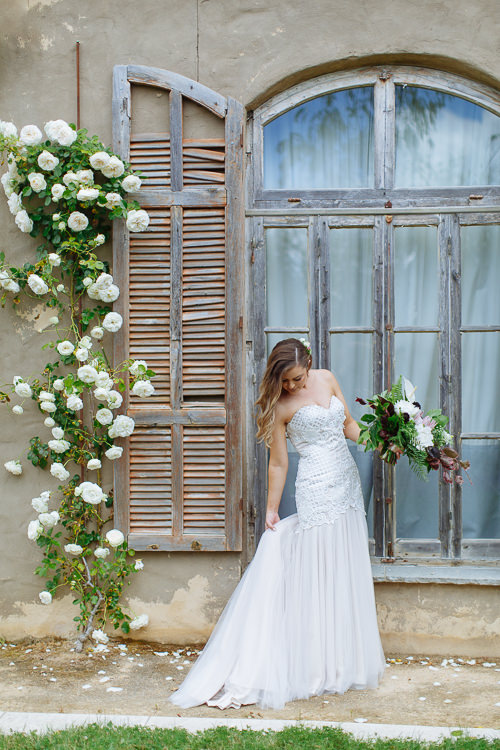 Wearing the strapless beaded gown, Elysha looks down at her bouquet beside the shuttered window.
