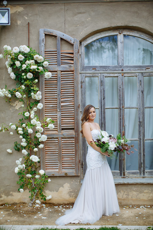 Elysha smiles at camera holding her protea and anthurium bouquet before the shuttered window.