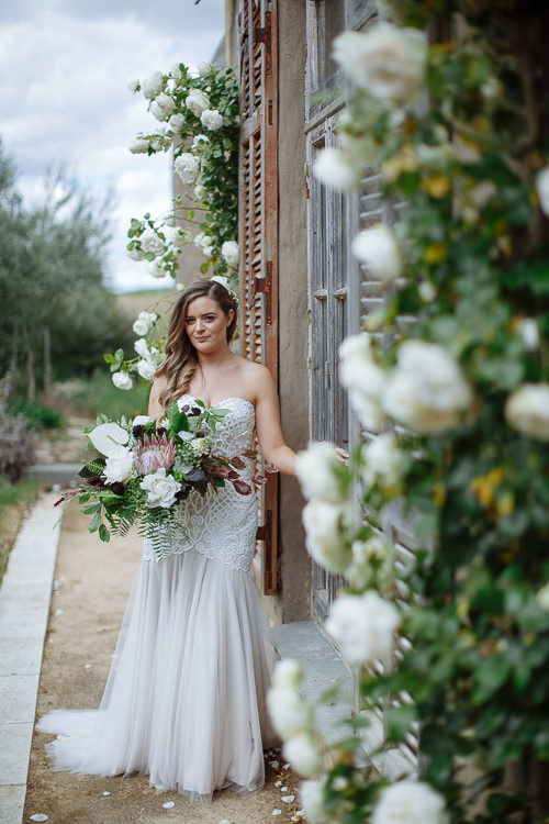 Through a foreground of white roses, Elysha smiles with her bouquet at a shuttered door.