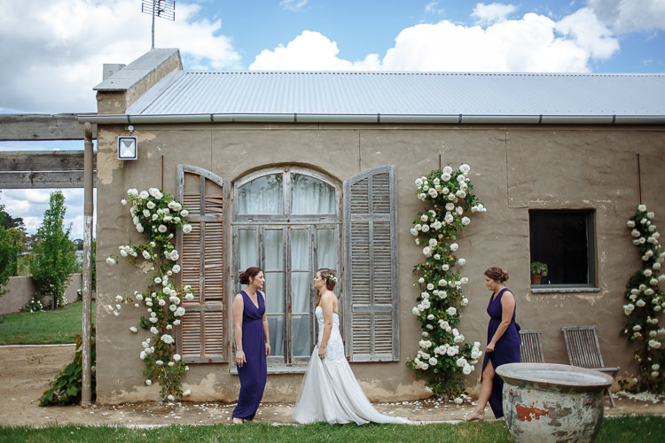 Elysha crosses the lawn between two bridesmaids in navy before the rose framed window.
