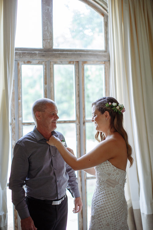 At the arched window, Elysha pins a white boutonniere to a bald man's charcoal shirt.