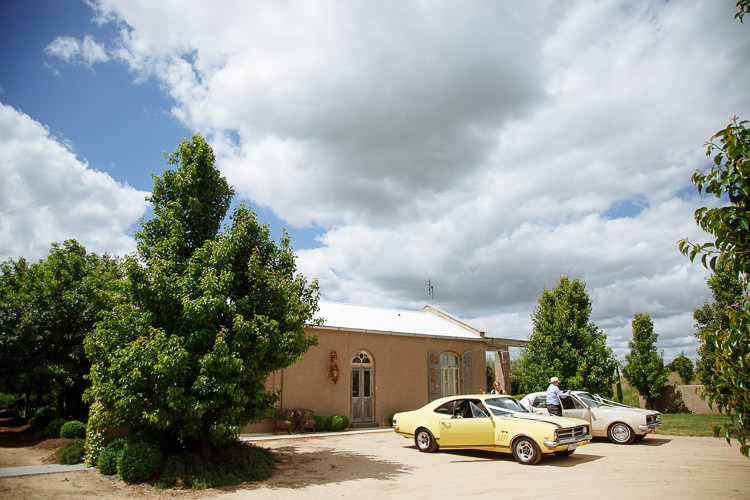 A yellow and a cream vintage sedan wait on the gravel before the rendered building.