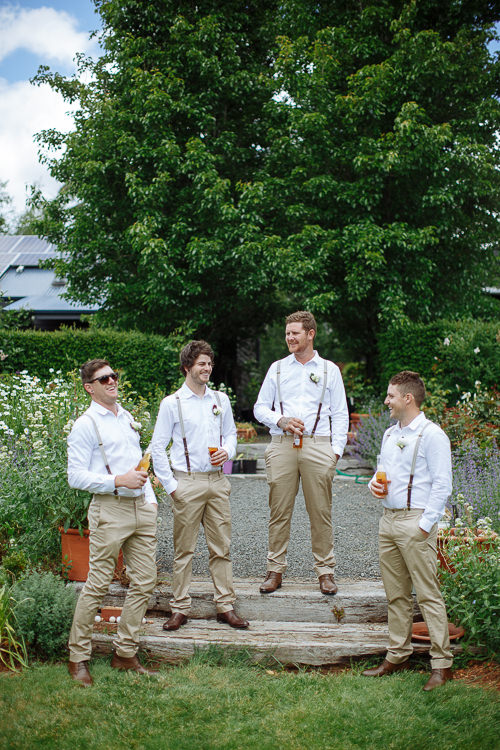 Four men in white shirts, braces and tan chinos laugh with beers on the garden steps.