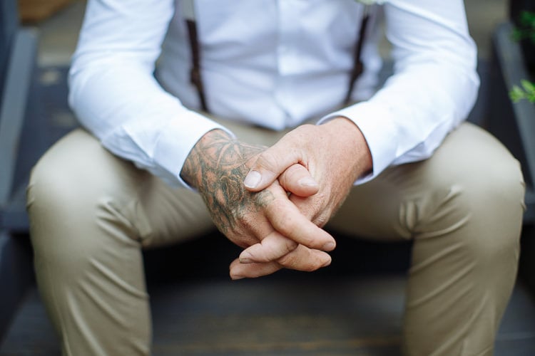 Close on clasped tattooed hands between knees in tan chinos, brace clips at the waist.