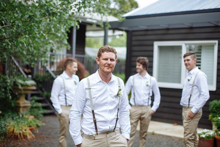 Mitch looks at camera with three groomsmen soft behind him against the black weatherboard building.