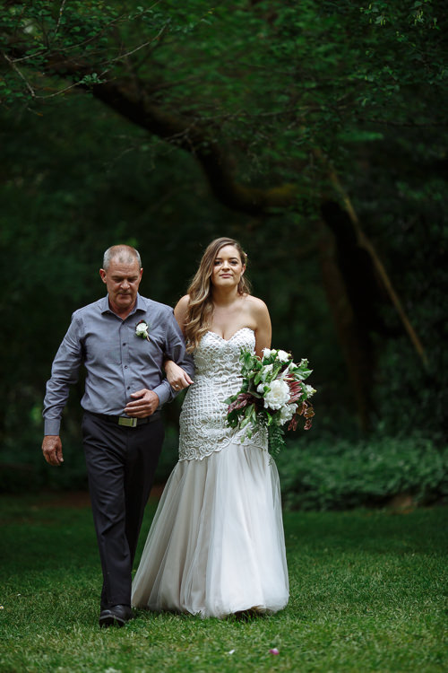 On the arm of a bald man in charcoal, Elysha crosses the lawn beneath a big tree.