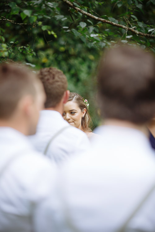 Between the blurred heads of the groomsmen, Elysha looks toward Mitch with flowers in her hair.